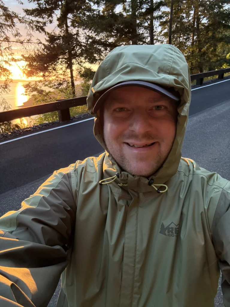 Robert Fleming outdoors in a green rain jacket, with trees and sunset over the water behind him